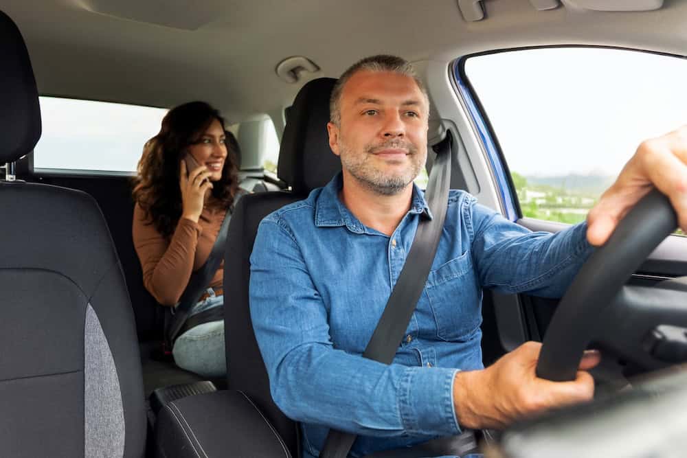Man driving while a woman sits in the back seat talking on the phone
