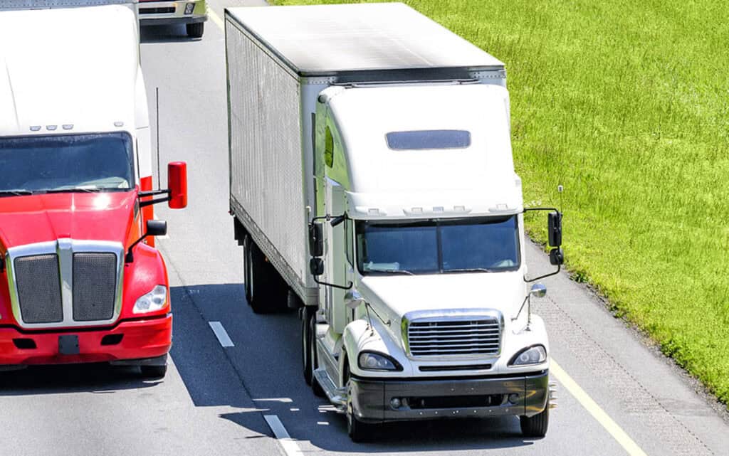 A red semi-trailer and a white semi-trailer driving down the road with the white semi ahead of the red.