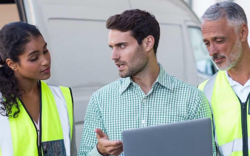 A man explaining information on a computer screen to two people on both sides of him in safety vests.