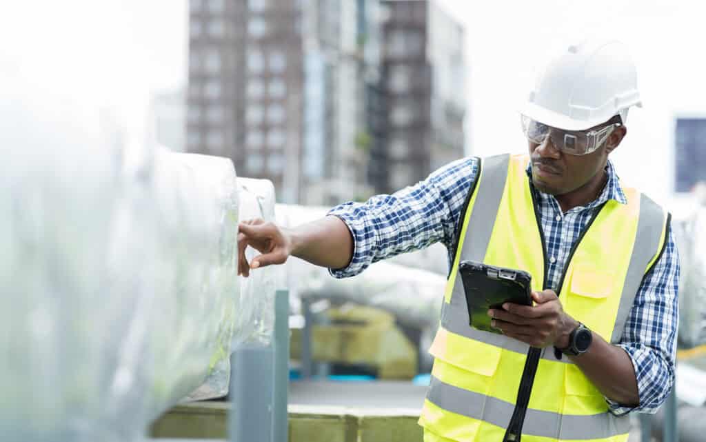 Man in hardhat and yellow vest inspecting an insulated pipe with a tablet in his hand